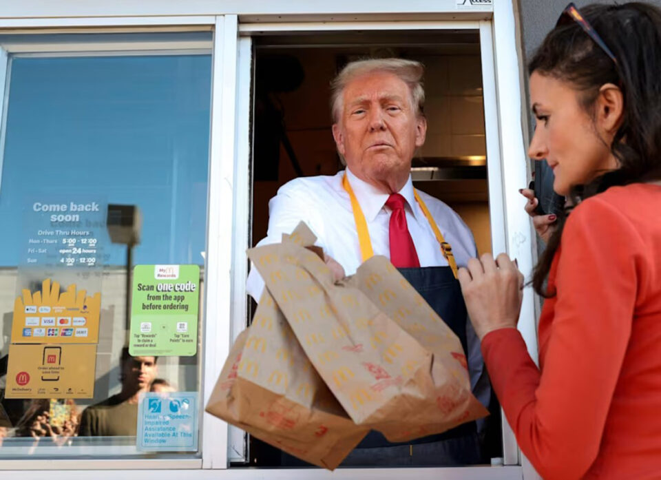 Donald Trump ojentaa pikaruokapusseja pikaruokakioskin ikkunasta. Edustalla tummatukkainen, punaisiin pukeutunut nainen ikään kuin vastaanottamassa annoksia.