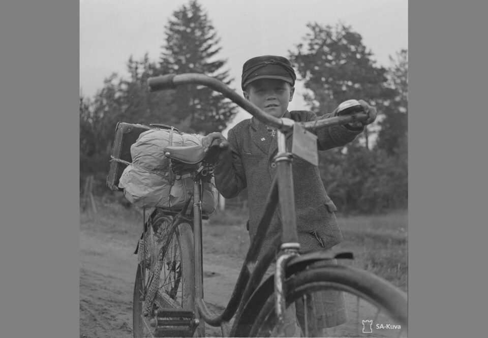 Vanhassa mustavalkokuvassa pieni poika taluttaa isoa polkupyörää, jonka tarakalla on paketti.