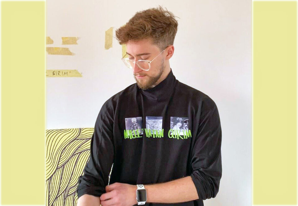 Silmälasipäinen nuori mies yllään musta, pitkähihainen paita, jonka rintamuksessa lukee: "Millet, Vatam, Qirim".