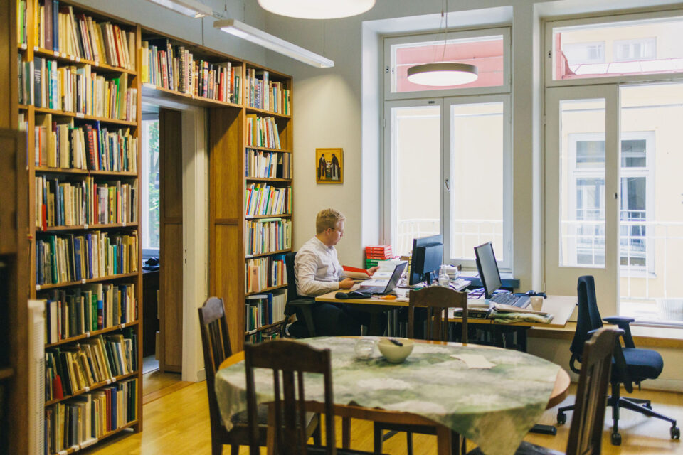 Nuori mies istuu tietokoneen ja työpöydän ääressä, vasemmalla puolellaan on suuret, korkeat ikkunat, takanaan seinää peittävät kirjahyllyt täynnä kirjallisuutta.