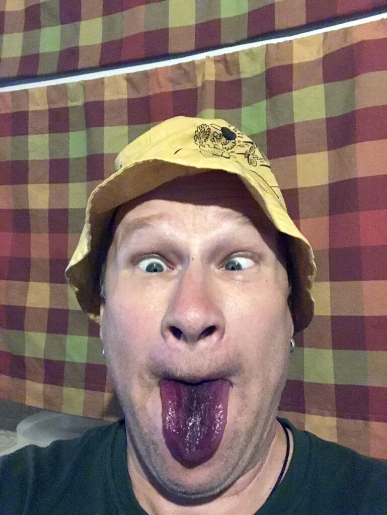 Lähikuvassa mies keltaisessa kalastajanlakissa, virnistää mustikan sinistämä kieli pitkällä ulkona suusta.