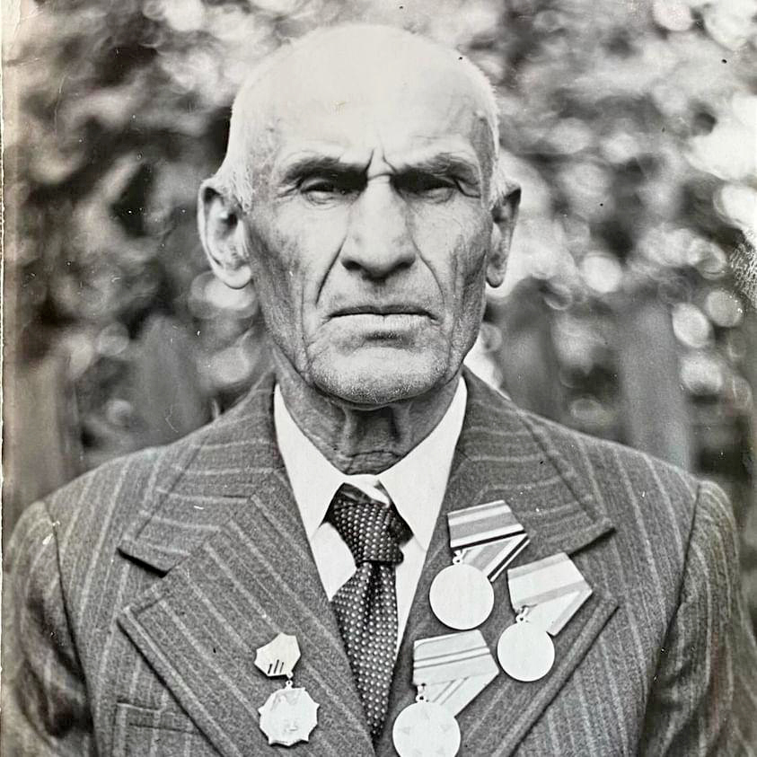 Mustavalkokuvassa tuiman näköinen iäkäs mies pukeutuneena harmaaraidalliseen puvuntakkiin ja tummaan kravattiin. Takin kauluksiin on kiinnitetty koko joukko kunniamerkkejä.