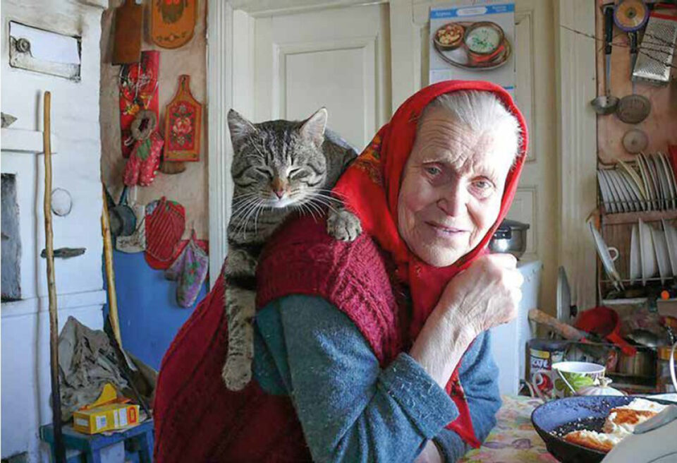 Iäkäs, punahuivinen nainen istuu pöydän ääressä etukumarassa hymyillen, olkapäällään loikoilee tyytyväinen harmaaraidallinen kissa.