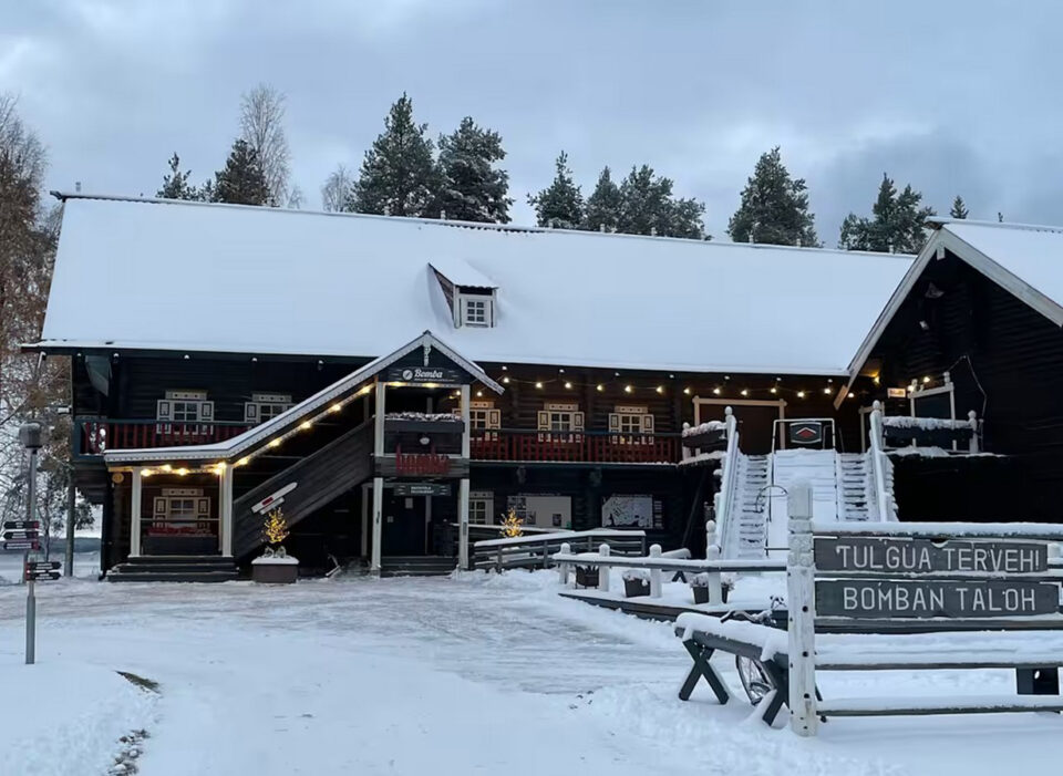 Lumisessa maisemassa vanha karjalaistyylinen, iso hirsirakennus, jonka vierellä olevassa kyltissä lukee karjalan kielellä: "Tulgua terveh Bomban taloh!"
