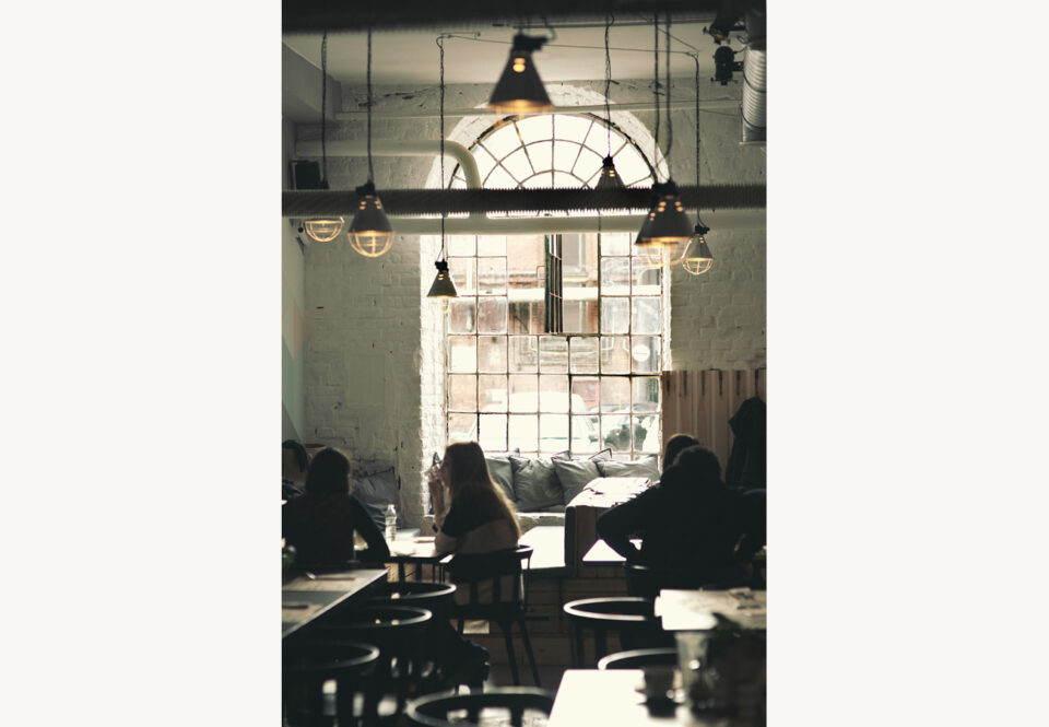 Nuoria ihmisiä kahvilapöytien äärellä. Taustalla on korkea, suuri ikkuna, josta tulvii runsaasti valoa sisään kahvilaan.