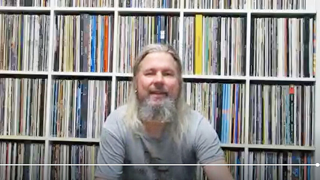 Parrakas, pitkähiuksinen, keski-ikäinen mies katsoo hymyillen kameraan, taustalla on iso hylly täynnä satoja vinyylilevyjä.