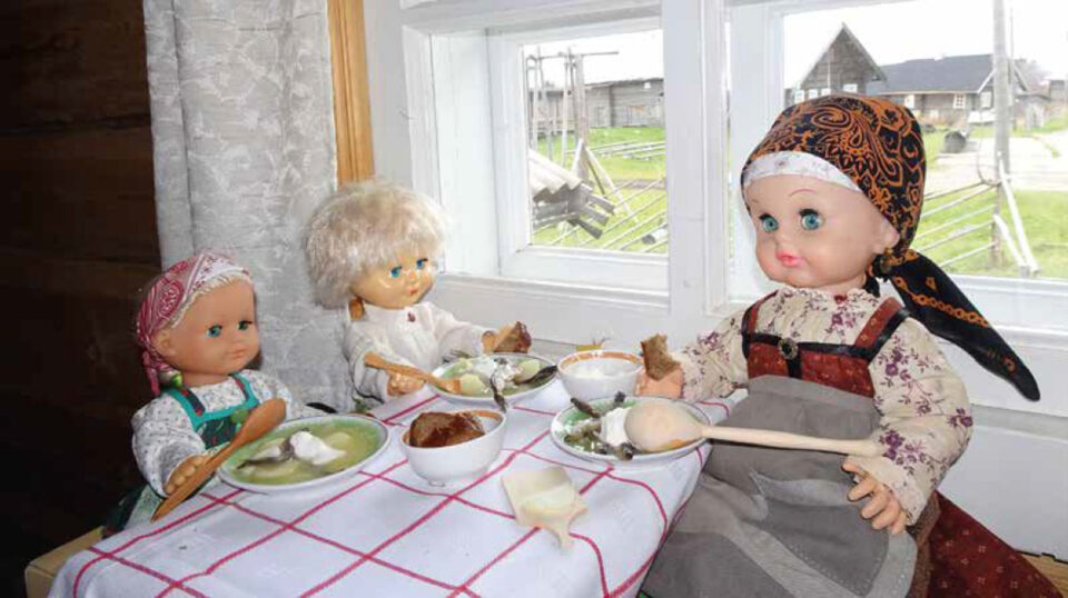 Karjalaisiin kansanpukuihin puetut nuket pienen pirtinpöydän äärellä, pienen "kalakeittoaterian" äärellä.