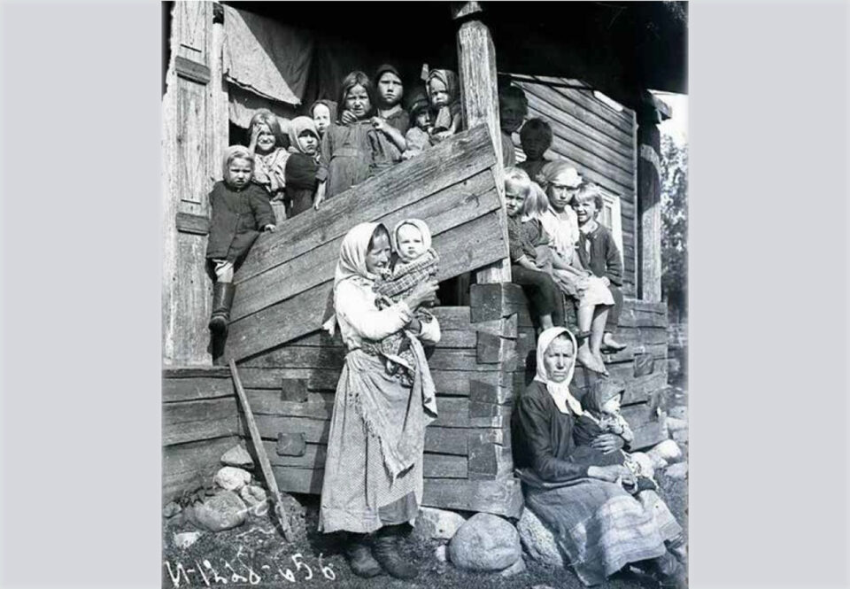 Vanhassa mustavalkokuvassa maalaamattoman hirsirakennuksen kuistilla seisoskelee tiiviisti kylki kyljessä 13 lasta. Kuistin edustalla istuu iäkäs nainen yksi pieni lapsi sylissään, lähempänä kameraa kuistin edustalla seisoo nuorempi nainen pienoikainen sylissään.