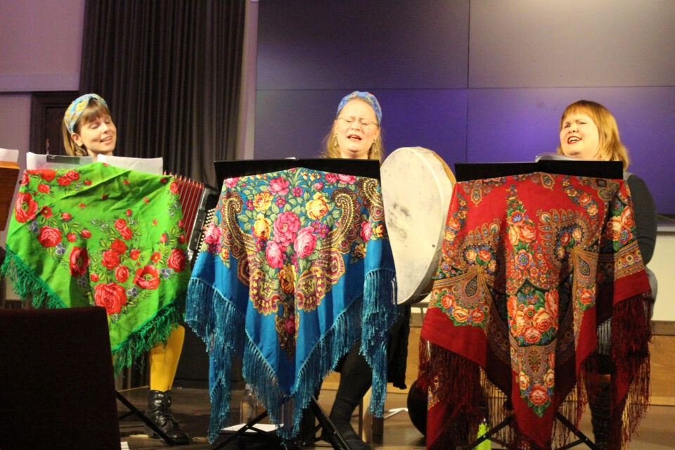 Kolme naista istuu tuoleillaan esiintymislavalla soittaen ja laulaen. Jokaisen edessä on pöytätaso, jota peittää värikäs, hapsullinen ruusuhuivi.