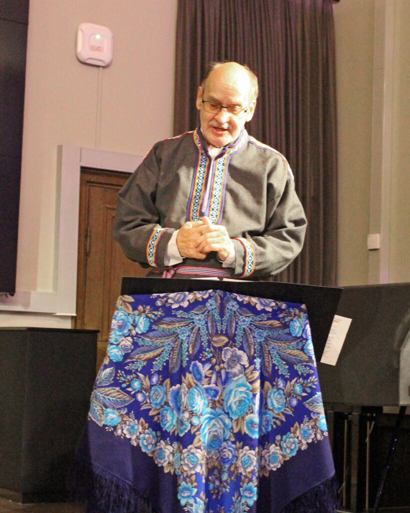 Iäkäs mies harmaasävyisessä saamelaismiehen kansanpuvussa laulaa kädet yhteen liitettyinä, katse on luotu alas pöytätasolle, jota peittää sinisävyinen kuviollinen hapsuhuivi.