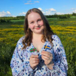 Pitkähiuksinen nuori nainen kesäisessä maisemassa, kukkamekossaan hymyilee iloisesti kohti kameraa. Kummassakin kädessä on pieni lippu: Itä-Karjalan ja Ruotsin liput.