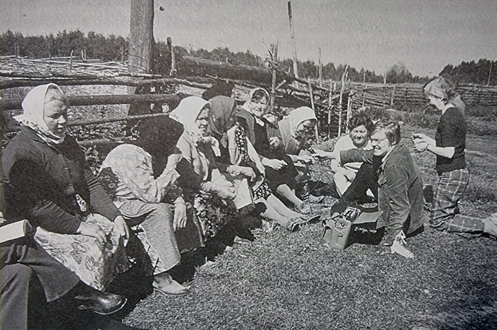 Mustavalkokuvassa maalaismaisemassa, maassa istuvia ihmisiä, joukko huivipäisiä naisia ja heitä vastapäätä kolme hymyilevää paljaspäistä ihmistä.