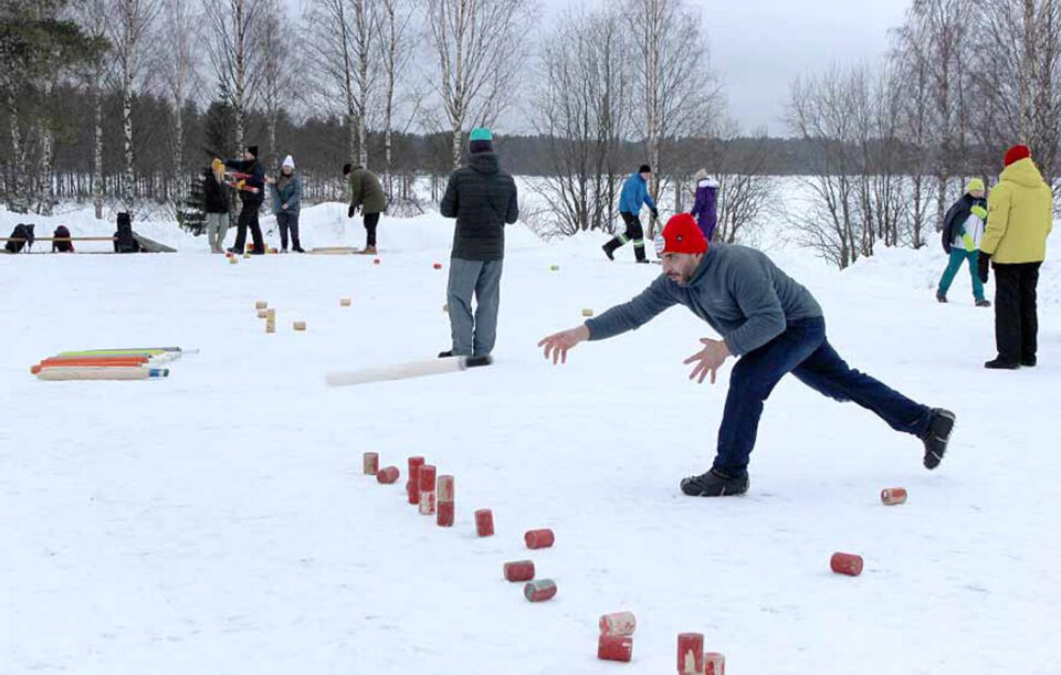Lumisella kentällä maassa kyykkäpelin palikoita, punapipoinen mies heittää kyykkämailaa, ympärillä ihmisiä talvivaatteissaan.