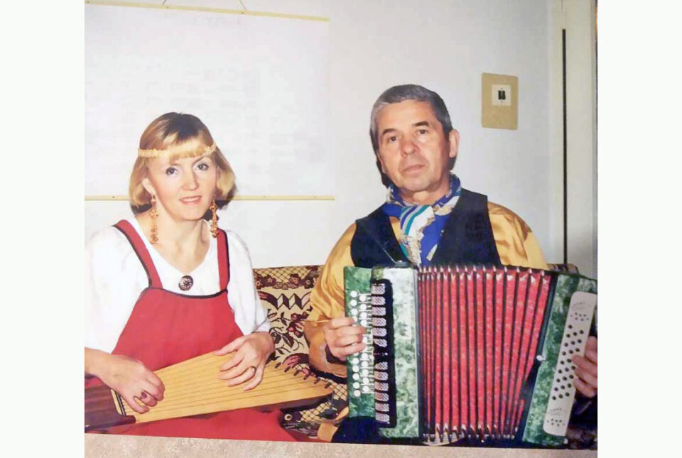 Karjalaisiin kansanpukuihin pukeutuneet nainen ja mies sylissään soittimet: naisella viisikielinen kantele, miehellä haitari.