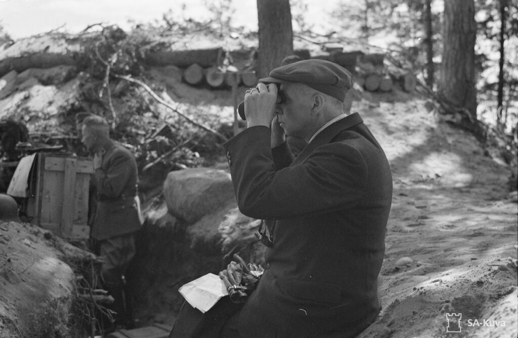 Juoksuhaudalta näyttävässä metsämaastossa tummaan puvuntakkiin ja lyhytlippaiseen lakkiin sonnustautunut mies kiikaroi vasemmalle päin. Taustalla näkyy toinen mies puulaatikkoa ojan tai juoksuhaudan pohjalla kantaen. Mustavalkoinen valokuva.