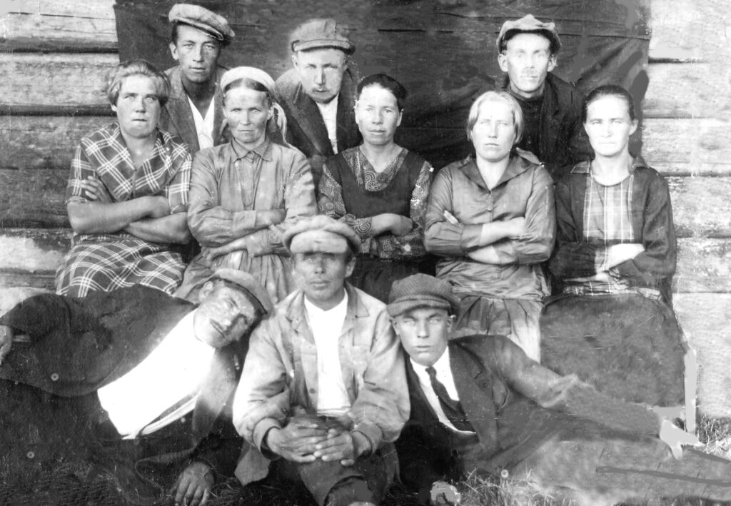 Vanha mustavalkoinen, hieman ylivalottunut kuva, jossa kameraa kohti katsoo 11 arkisiin vaatteisiin sonnustautunutta miestä ja naista: eturivissä kolme miestä maassa istuen tai toisiinsa nojaillen, takarivissä kolme miestä, heidän välissään ehkä portaalla istuen neljä naista. Kaikilla on totiset ilmeet.