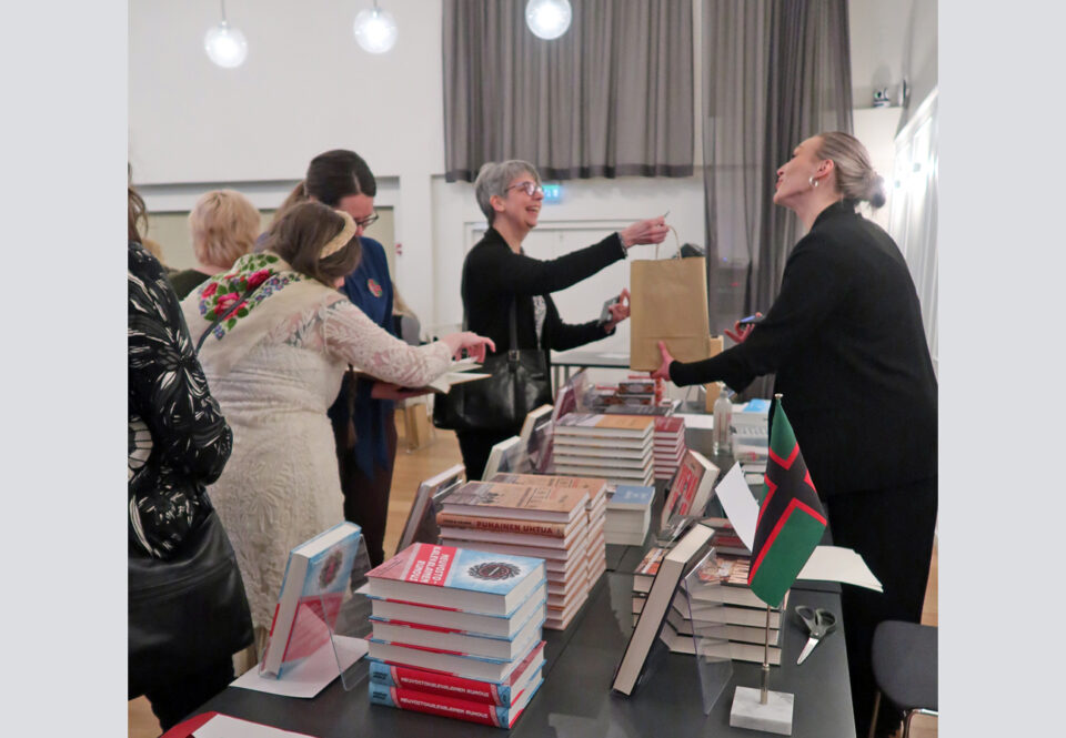 Pitkä myyntitiski täynnä kirjoja, etualalla Itä-Karjalan puna-vihreä-musta lippu, tiskin takaa myyjä ojentaa ruskeaa paperikassia hymyilevälle ostajanaiselle, kolme-neljä ihmistä katselee myyntipöydällä olevia kirjapinoja.