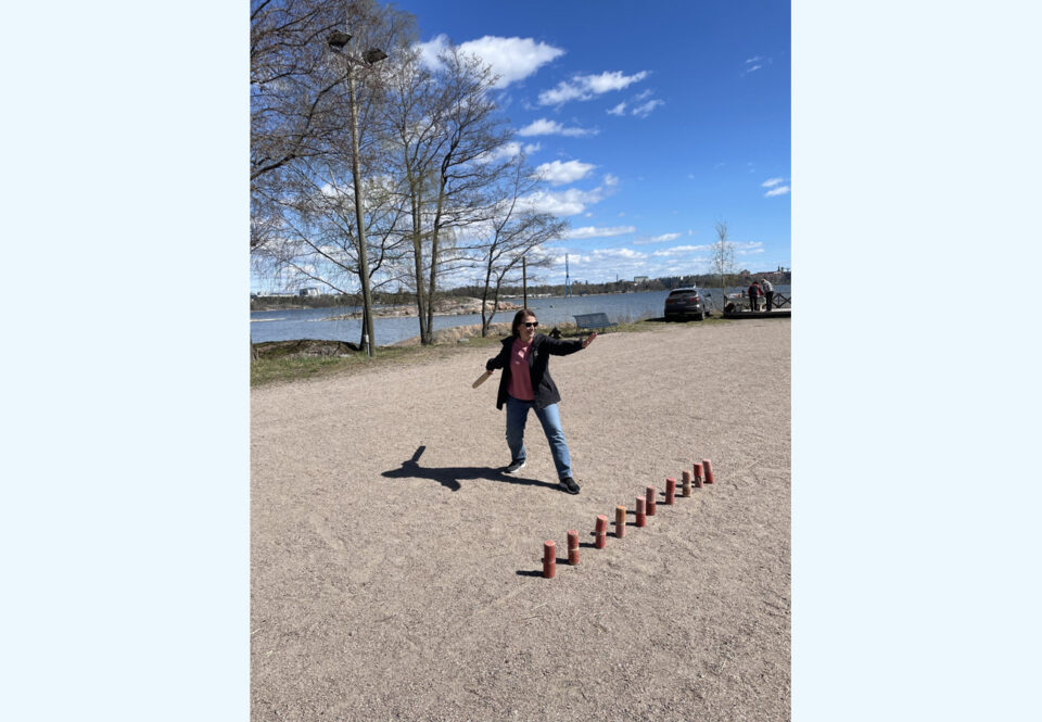 Hiekkakentällä on maassa rivi puupalkkeja, niiden edessä ulkoiluvaatteisiin pukeutunut nainen on valmiina heittämään kyykkämailaa. Taustalla on keväinen maisema, puissa ei ole vielä lehtiä, mutta taivas on hyvin kirkkaansininen ja vain muutamia pieniä poutapilviä on näkyvissä.