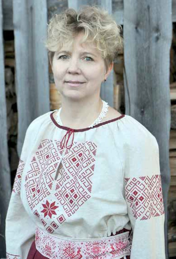 Vaaleatukkainen nainen katsoo kameraan puolilähikuvassa. Hänellä on yllään kaunis paita ja leveä kankainen vyö, joissa on runsaita punapoimintakirjailuja, eli punaisella langalla valkoiselle pohjalle kirjottuja perinteisiä, karjalaistyylisiä kuvioita.