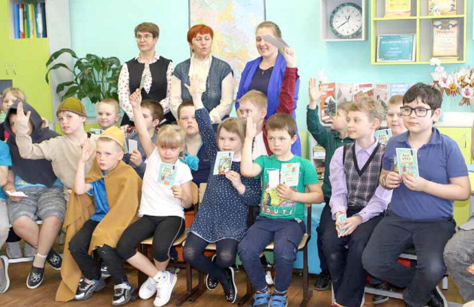 Ryhmä lapsia istuu tuoleilla, osa heistä viittaa ja esittelee käsinpiirrettyjä kortteja. Lasten takana seisoo kolme aikuista naista.