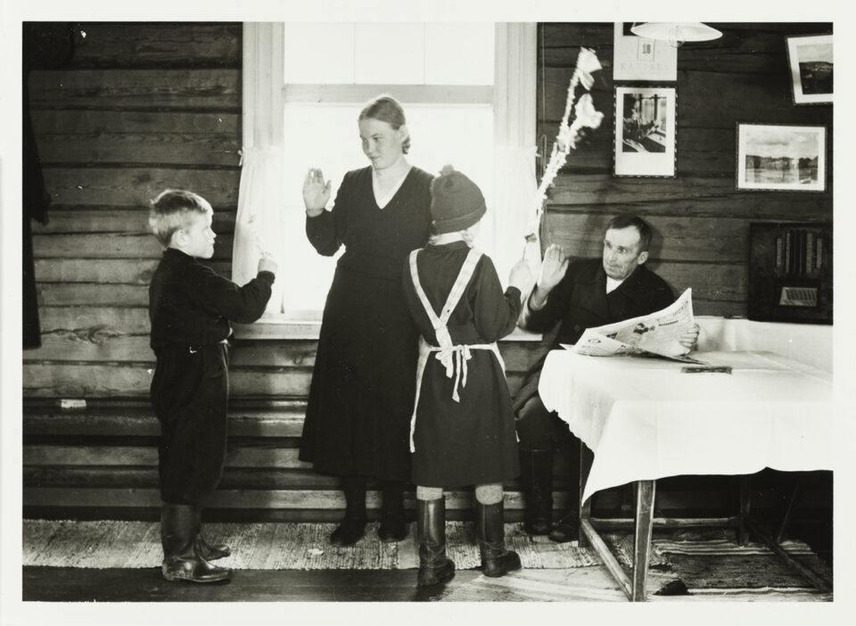 Mustavalkoinen valokuva, jossa hirsiseinäisen tuvan penkillä istuu mies, ikkunan ääressä seisoo nainen. Kaksi lasta virpoo häntä parhaillaan.