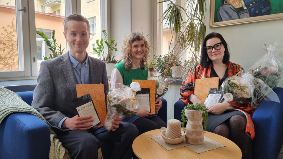 Kolme nuorta ihmistä istuu kirkkaansinisellä sohvalla valoisassa ikkunasyvennyksessä. Kaikilla on kädessään teepakkaus, kunniakirja ja kukkakimppu.