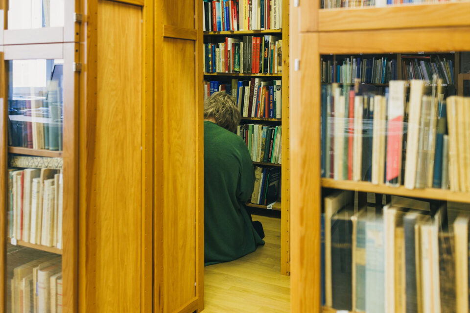 Puisten, kirjoja täynnä olevien hyllyjen keskellä lattialla istuu vihreään villatakkiin pukeutunut nuori ihminen selin, kumartuneena tutkimaan kirjaa tms.