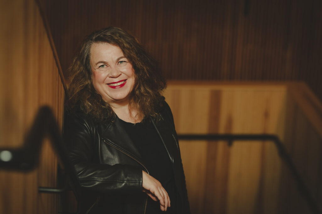 Leveästi hymyilevä, punahuulinen, mustaan nahkatakkiin pukeutunut nainen seisoo portaikossa, porraskaiteeseen nojaten ja katsoo ylöspäin kohti kuvaajaa.