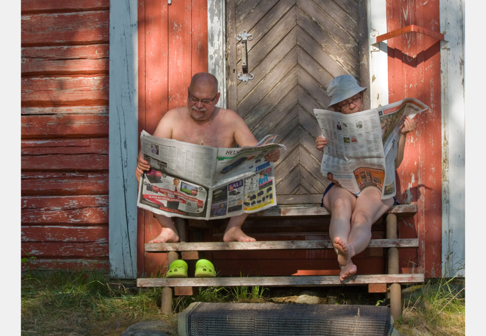 Punamullalla maalatun kesämökin rappusilla istuvat mies ja nainen helleasuissaan, mies ilman paitaa, naisella yllään shortsit ja hellehattu. Kumpikin on syventynyt lukemaan omaa sanomalehteään.