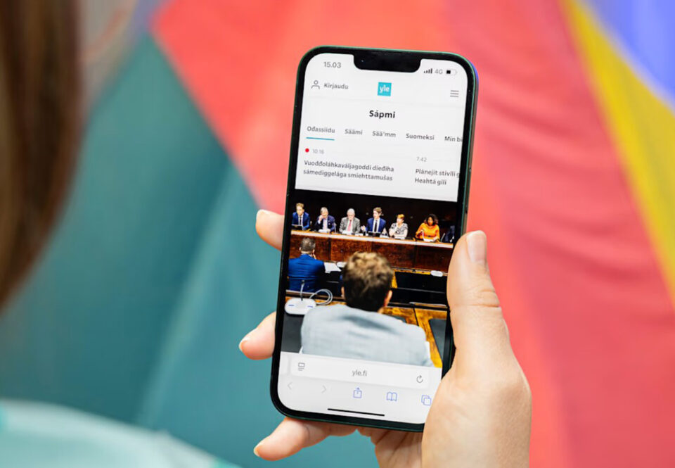 Olan yli otettu lähikuva, jossa näkyy matkapuhelimen näyttö. Näytöllä saamenkielistä uutistekstiä ja kuvaa eduskunnasta.