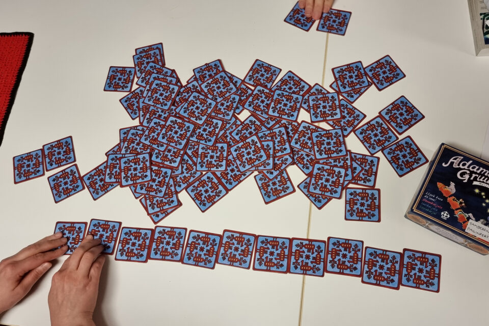 Valkoiselle pöytälevylle on levitetty värikkäitä muistipelikortteja, kuvapuoli alaspäin. Kahta puolen kädet sekoittelevat tai järjestelevät kortteja.