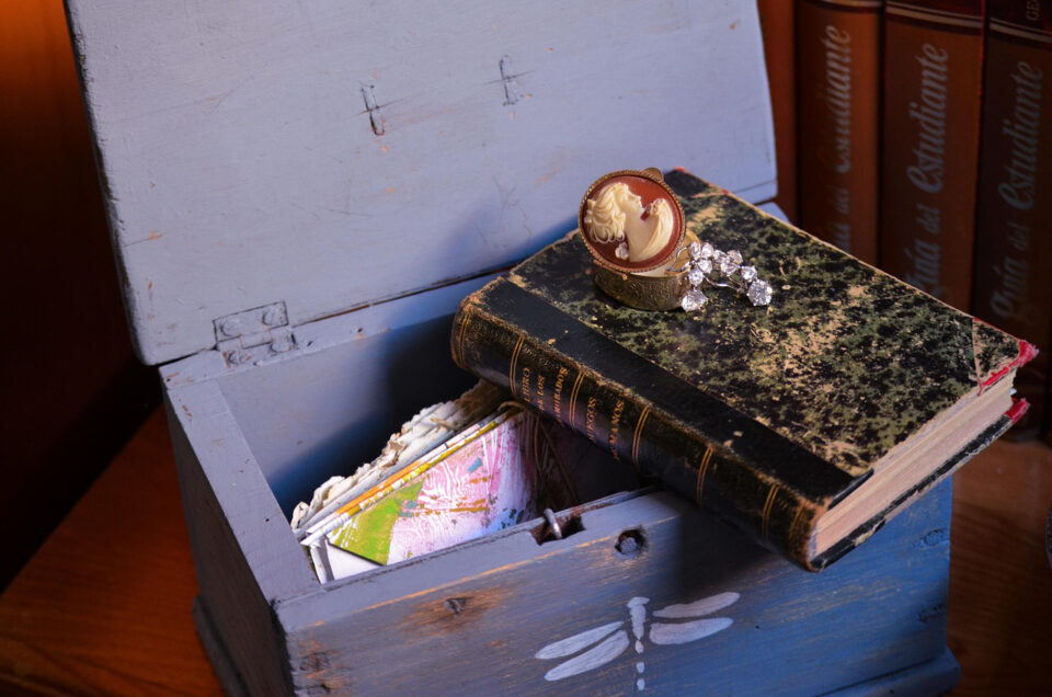 Siniseksi maalattu puinen korurasia, jonka kansi on auki. Sen sisältä pilkottaa kirjenippu, päällä on vanha mustakantinen kirja ja sen päällä vielä pieni Camee-kantinen korurasia.