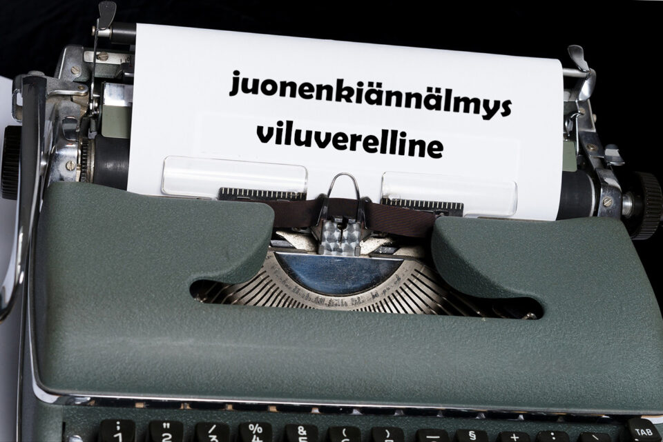 Mekaaninen, harmaa kirjoituskone, jonka telalle kierretyssä valkoisessa paperissa lukee allekkain kaksi karjalankielistä sanaa: "juonenkiännälmys" ja "viluverelline"