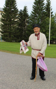 Karjalaiseen kansanpukuun pukeutunut iäkäs mies pitää toisessa kädessään vaaleapohjaista kukkahuivia, toisessa kädessä on vaaleanpunainen muovikassi ja puinen kävelykeppi. Taustalla näkyy kuusia ja lipputanko, mies seisoo hiekkatiellä hymyssä suin.