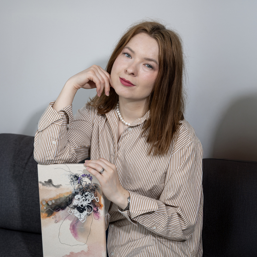Nainen istuu pöydän ääressä, nojaa oikealla kädellä kevyesti poskeensa, vasemman käden tuntumassa on akvarellimaalaus. Naisella on yllään beige paitapusero, ruskeat hiukset ulottuvat olkapäille, taustalla on hillityn harmaa seinä.