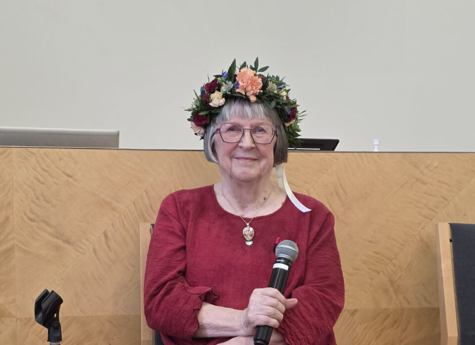 Harmaahiuksinen, silmälasipäinen nainen istuu mikrofoni kädessään ja hymyilee kameralle. Yllään hänellä on kirkkaanpunainen pellavamekko ja päässä muhkea kukkaseppele, jossa erivärisiä, tummanpunaisia ja vaaleampia kukkia.
