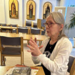 Harmaahiuksinen, silmälasipäinen nainen istuu pöydän ääressä. Pöydällä on lehtiä, huoneessa näkyy muitakin pöytiä, joiden ääressä kangaspäällysteisiä, selkänojallisisa tuoleja. Taustalla näkyy valkoinen seinä, jolla on kolme yläreunastaan kaarevaa, ikonin tyylistä maalausta. Jokaisessa maalauksessa on kuvattuna kullankeltaisella pohjalla ortodokisen kirkon pyhä henkilö.