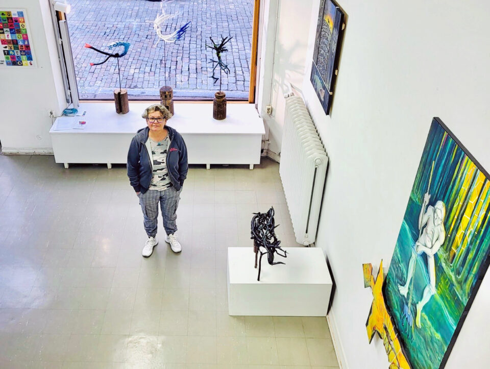 Lintuperspektiivistä kuvattu taidegalleriatila, jossa ihminen seisoo kädet taskuissa suuren ikkunan edessä. Ikkunalaudalla on veistoksia, seinillä maalauksia. Ihminen on pukeutunut rentoon huppariin,harmaisiin housuihin ja valkoisiin lenkkitossuihin.