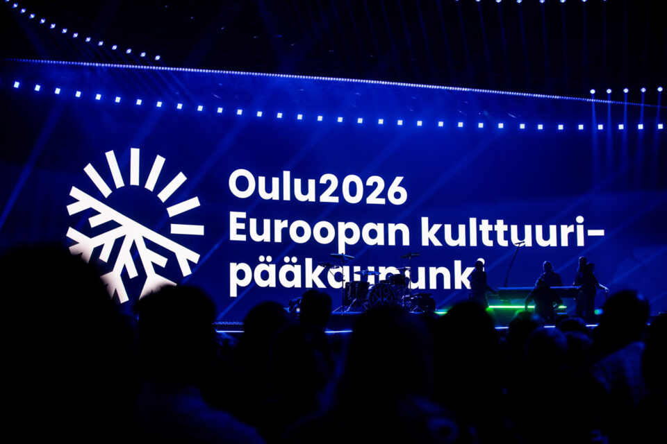 Suuri seinälle heijastettu sinipohjainen "lakana" tai banneri, jossa valkoisella teksti "Oulu2026 kulttuuripääkaupunki" ja tapahtumatunnus. Bannerin edustalla näkyy yleisöä silhuetteina, mustina varjokuvina.