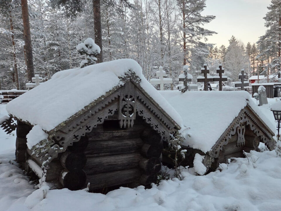 Pieniä hirsimökkimäisiä, matalia rakennuksia paksun lumipeitteen ympäröiminä. Rakennelmien katoilla on paksu lumikerros, kattoja ympäröivät koristeleikkauksin veistetyt laudat.