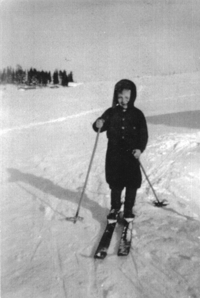 Pieni poika hiihtää leveilä puisilla suksilla lumisessa maisemassa, hymyilee karvalakkinsa alta kohti kameraa. Yllä on tumma pusakka ja pussihousut, sauvat ova pitkähköt ja porkat isot ja pyöreät.