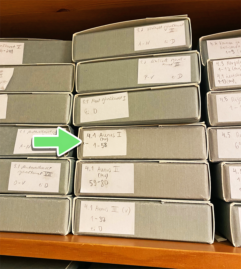Puisen, kirsikkasävyisen hyllyn päällä on kerroksittain harmaita arkistolaatikoita. Yhtä arkistolaatikkoa kohti kuvaan on jälkeenpäin lisätty vihreä korostusnuoli osoittamaan arkistolaatikkoon liimattua tarralappua, jossa lukee: "4.1 Aunus I (mv) 1 - 58"