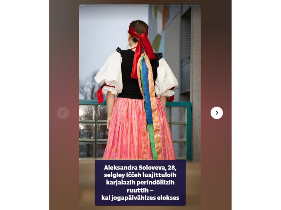 Nuori nainen seisoo selin kameraan yllään värikäs karjalainen kansanpuku: vaaleanpunainen raidoitettu pitkä hame, valkoinen pussihihainen paita, musta liivi, hiukset on sidottu pitkillä värikkäillä nauhoilla, pään ympäri kiertää leveä ja pitkä punainen silkkinauha, myös paidan hihansuissa on samaa punaista kiiltävää nauhaa.