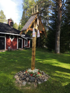 Puinen vienalainen, katettu matkamiehen risti seisoo viheriöivällä pihalla, taustalla punaiseksi maalattu puurakennus, oikealla puolella häämöttää iso mänty. Matkamiehen juurelle on aseteltu kauniisti keko kiviä. Ristin poikkipuulla lepää valkoisesta pellavasta tehty, punapoimintakirjailuin koristeltu karjalainen käspaikka.