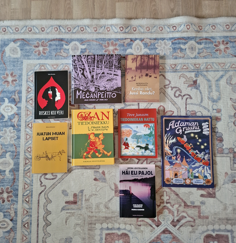 Lattialla, vaaleasävyisen, itämaisen kuviollisen villamaton päällä on kahdeksan karjalankielistä kirjaa etukansi ylöspäin.