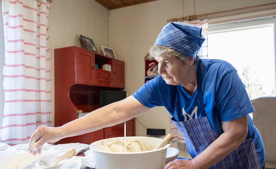 Siniseen huiviin ja esiliinaan sekä siniseen t-paitaan pukeutunut nainen levittää jauhoja leivinalustalle, vieressä on iso valkoinen taikinakulho täynnä kohonnutta taikinaa.