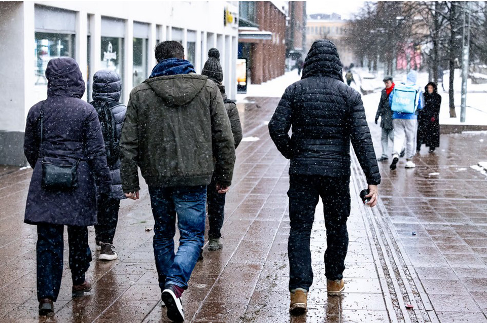 Tummiin, hupullisiin takkeihin ja housuihin pukeutuneet ihmiset kävelevät räntäsateisella kadulla selin kameraan.