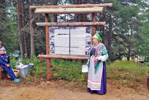 Nainen karjalaisessa perinneasussa seisoo puisen infokyltin vierellä