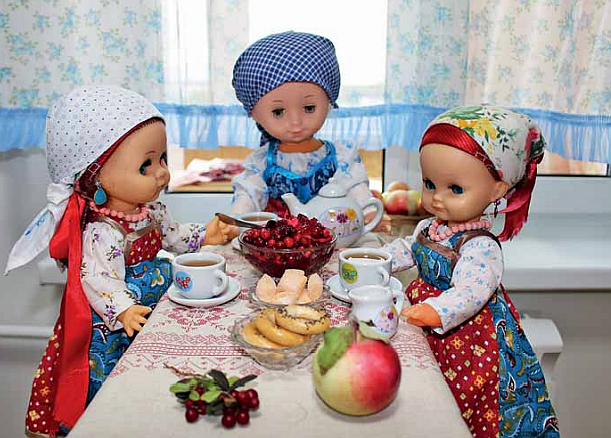 Kolme nukkea pöydän äärellä ja pöydässä on teetä, hedelmiä ja keksejä