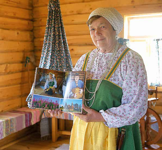 Hymyilevä iäkäs nainen pitelee käsissään kirjaa puutalon sisällä